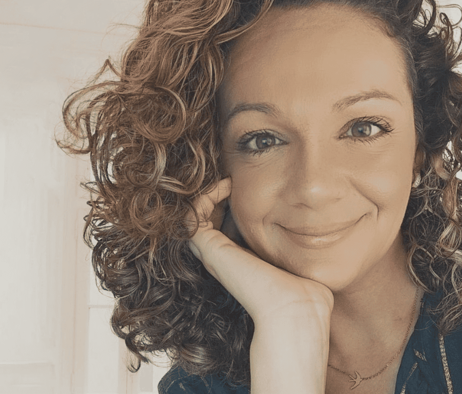 Mulher sorridente com cabelo cacheado castanho, apoiando o queixo na mão, transmitindo uma expressão acolhedora e profissional.